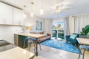 A modern kitchen with white cabinets, a dining area with a blue rug, and a balcony with a view of palm trees.