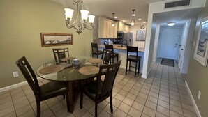 Un coin repas avec une table en verre entourée de chaises, un lustre et une cuisine en arrière-plan.