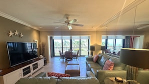 A spacious living room with a large flat-screen TV, comfortable seating, and a balcony with a view of palm trees.