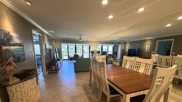 A spacious dining area with a large wooden table and white chairs, a kitchen in the background, and large windows providing natural light.