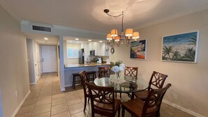 A dining area with a glass table surrounded by wooden chairs, a kitchen in the background, and a beach-themed painting on the wall. - Beachfront  2 bed/ 2 Bath Condo (Sarasota)