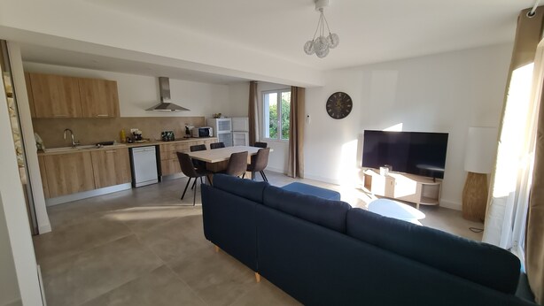 Una sala de estar moderna con un sofá azul oscuro, un comedor con mesa y sillas de madera, una cocina con gabinetes de madera y electrodomésticos blancos, y un televisor sobre un mueble.