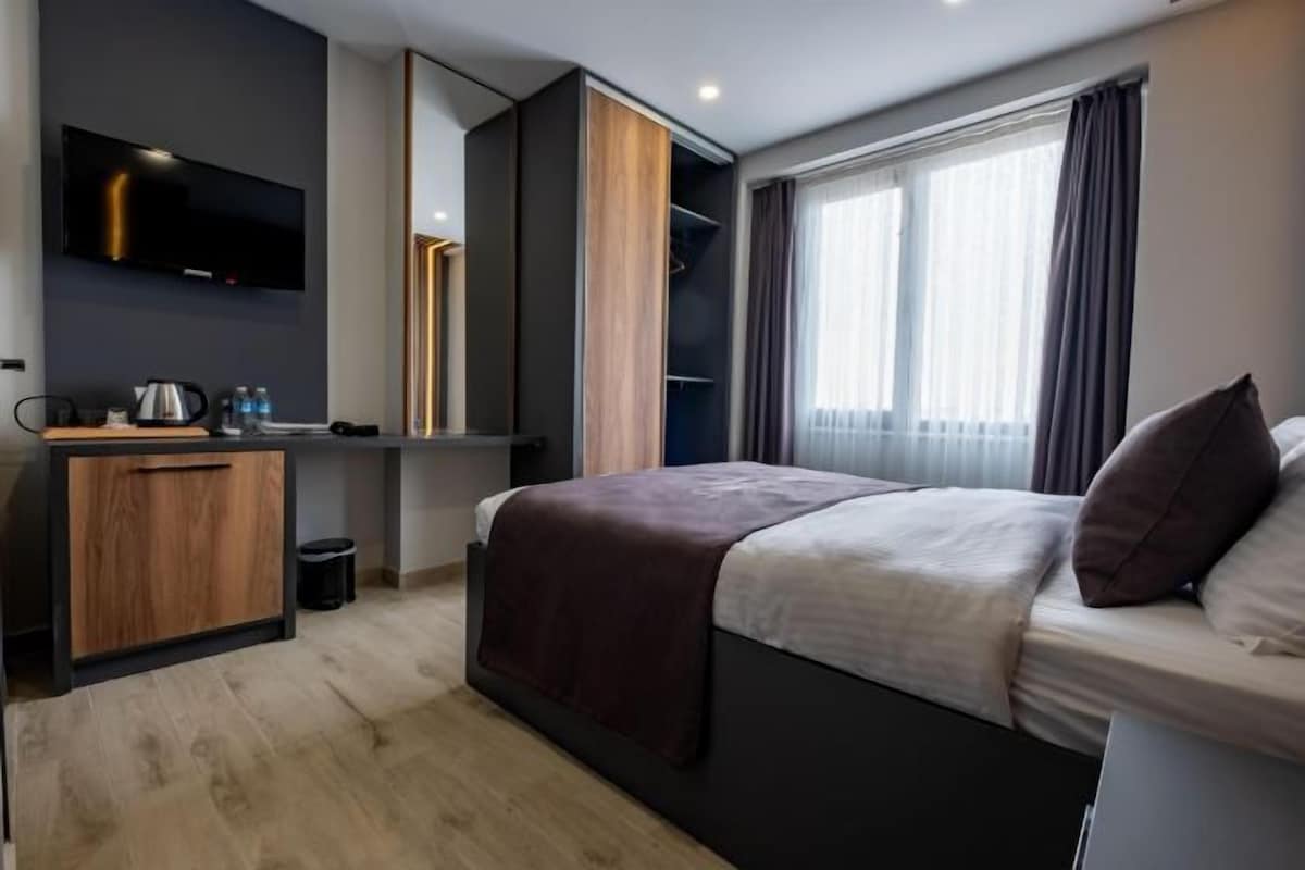 Ett modernt hotellrum med en säng, ett skrivbord, en TV och ett stort fönster med gardiner.