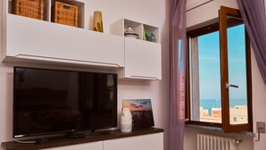 Un salon moderne doté d’un téléviseur à écran plat, de meubles blancs, de paniers en osier, de vases décoratifs et offrant une vue sur la mer par une fenêtre ornée de rideaux violets.