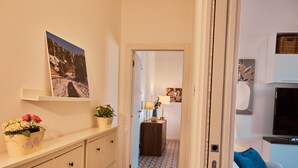 Un couloir avec un sol à motifs, un canapé agrémenté d’un coussin bleu, un téléviseur posé sur un meuble et une porte donnant sur une autre pièce.