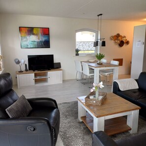 A modern living room with a flat-screen TV, a dining area with a table and chairs, a black leather sofa, and a large abstract painting on the wall.