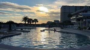 Una piscina con un atardecer de fondo, rodeada de palmeras y un edificio con balcones.