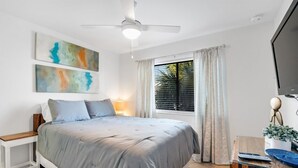 Un dormitorio con cama, ventilador de techo, ventana con cortinas y televisor de pantalla plana.