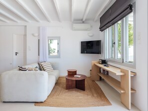 Una sala moderna con un sofá blanco, una mesa de centro de madera, un televisor montado en la pared y un ventanal con vistas a zonas verdes.