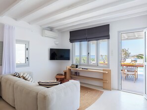 Un salon moderne avec un canapé, un téléviseur à écran plat, un bureau en bois et une vue sur la mer grâce à de grandes fenêtres.