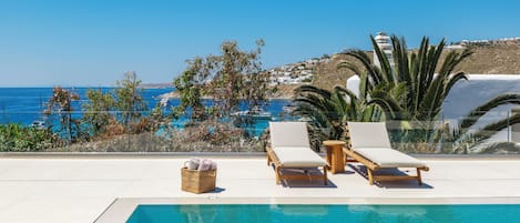Zona de piscina con sillones de descanso y una cesta de mimbre, con vistas a un paisaje costero con edificios y embarcaciones.