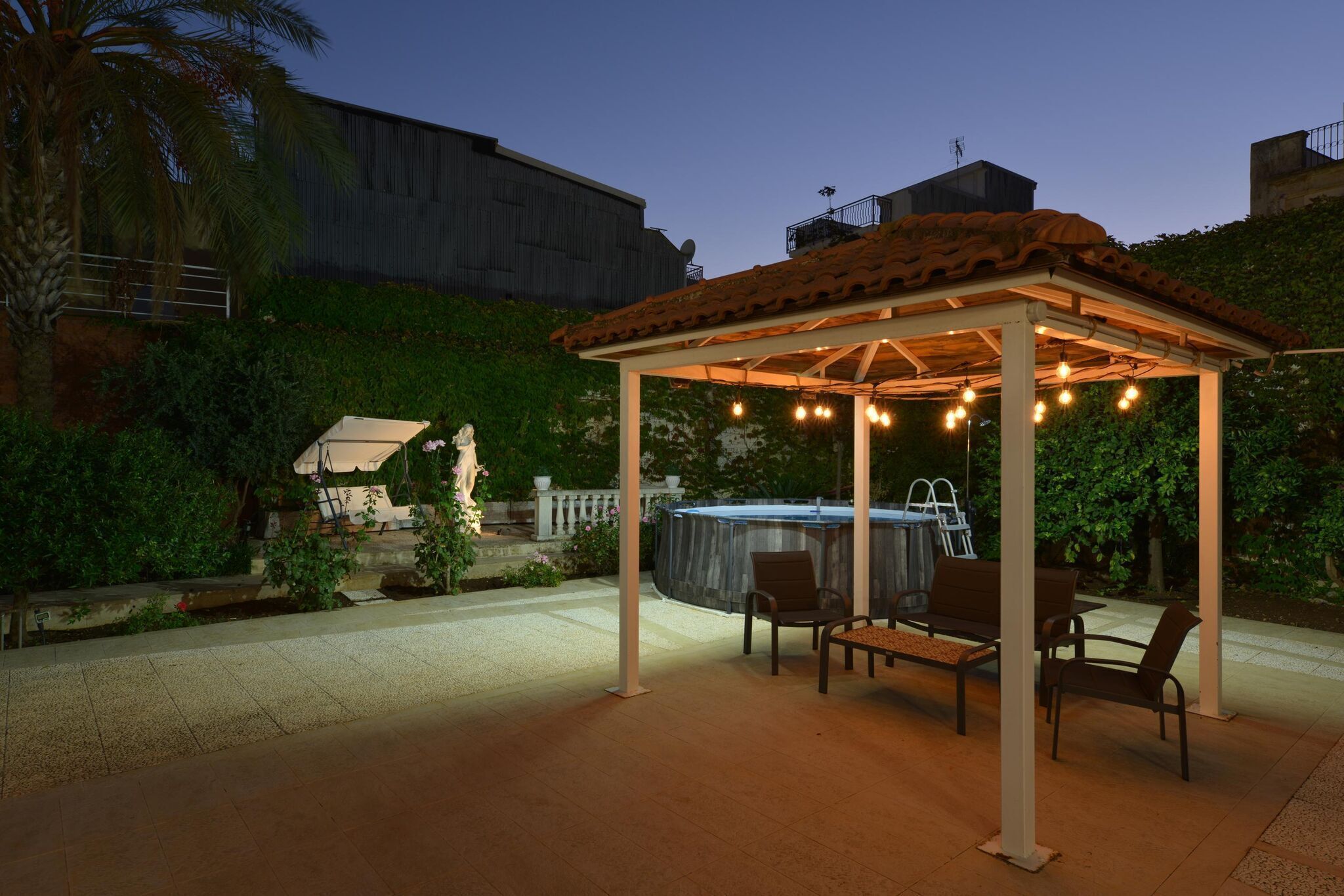 A covered outdoor seating area with a hot tub, surrounded by greenery and a building in the background.