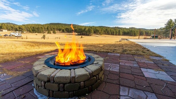 A fire pit with flames, surrounded by a brick walkway, with a forested hill in the background. - Custer Connection- Private Retreat: 6-Acre Nature Escape with Trail Access, Custer. (Custer)