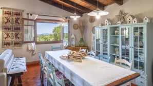A dining area with a table set for two, a window with a view of greenery, and a cabinet with glass doors. - Villa Mirasol with sea and panoramic view, ideal for 4 people with AC and WiFi (Sardegna)