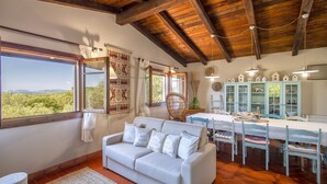 A cozy living room with a sofa, dining table, and chairs. There are windows with curtains, a wooden ceiling, and a view of greenery outside. - Villa Mirasol with sea and panoramic view, ideal for 4 people with AC and WiFi (Sardegna)