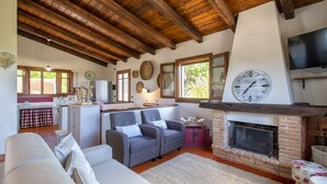 A cozy living room with a fireplace, wooden beams, and a kitchen area with a refrigerator and window. - Villa Mirasol with sea and panoramic view, ideal for 4 people with AC and WiFi (Sardegna)