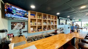 A bar area with a wooden counter, shelves with bottles, a TV displaying sports, and a sign about a lift. - Oceanfront Blockade Runner Villa - Pool (North Myrtle Beach)