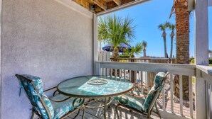 A covered outdoor patio with a round glass table and two chairs, a wooden ceiling, and a view of palm trees and a beach. - Oceanfront Blockade Runner Villa - Pool (North Myrtle Beach)