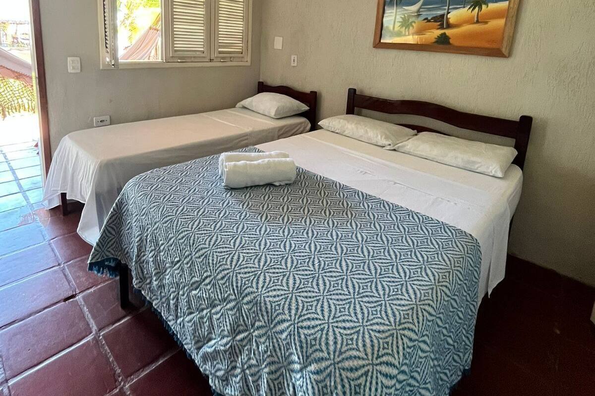 A hotel room with two single beds, a large bed with a patterned bedspread, a painting of a beach scene, and an air conditioning unit on the wall.