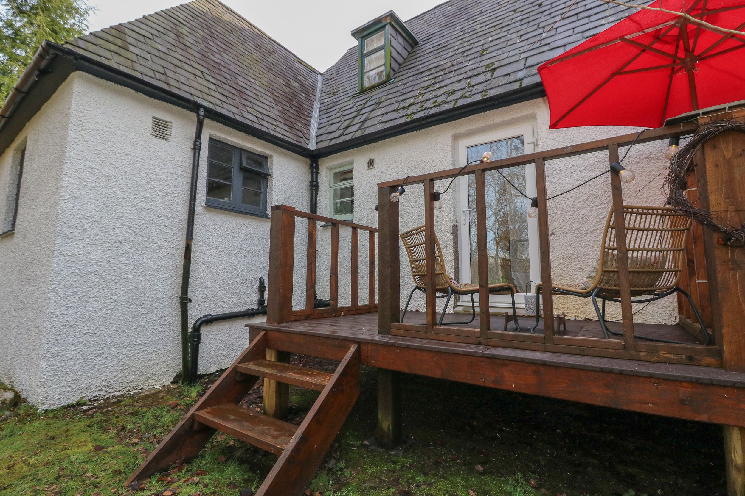 A wooden deck with chairs and a red umbrella, leading to a white building with a sloped roof.