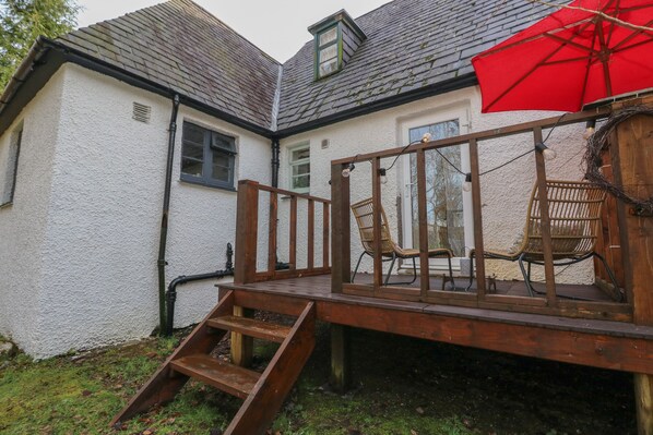 A wooden deck with chairs and a red umbrella, leading to a white building with a sloped roof. - The Cottage In The Woods (Taynuilt)