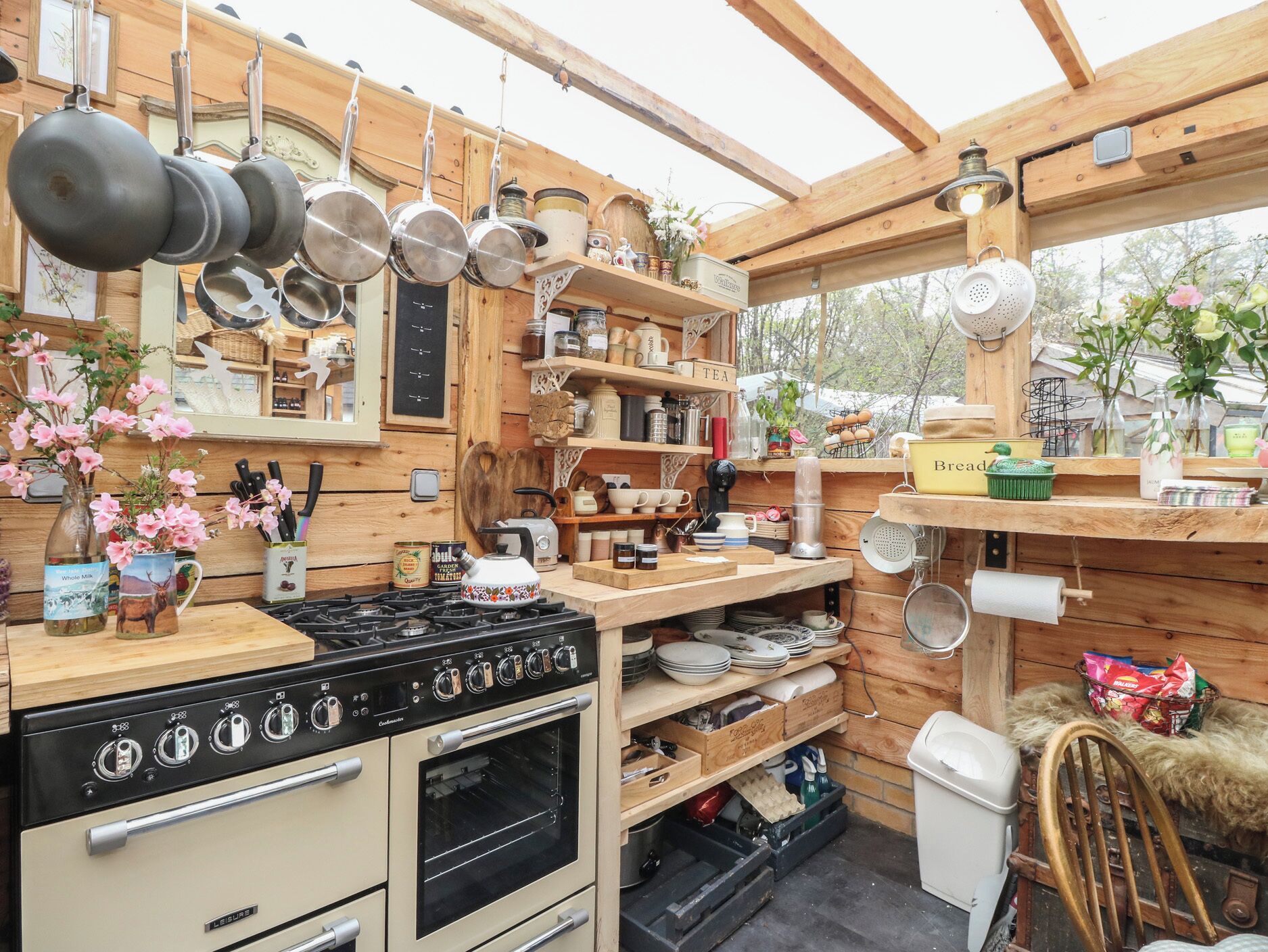 A rustic kitchen with a stove, oven, hanging pots, and wooden shelves with various kitchen items.