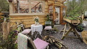 A wooden cabin with a window, a small table with chairs, and a garden area with plants and a fire pit. - The Cottage In The Woods (Taynuilt)