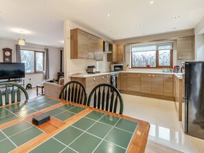 A modern kitchen with wooden cabinets, a dining table with chairs, a TV, and a window with a view of trees.