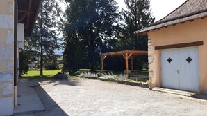 A covered outdoor seating area with a pergola, a gravel driveway, and a building with a garage. - Maison Indépendante en Chartreuse (Saint-Laurent-du-Pont)