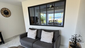 A modern living room with a grey sofa, a large window with a view, a ceiling fan, and a decorative tree sculpture.