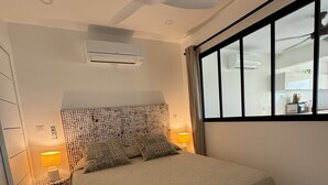 A bedroom with a bed, a ceiling fan, an air conditioner, a bedside lamp, a window, and a view of the kitchen.