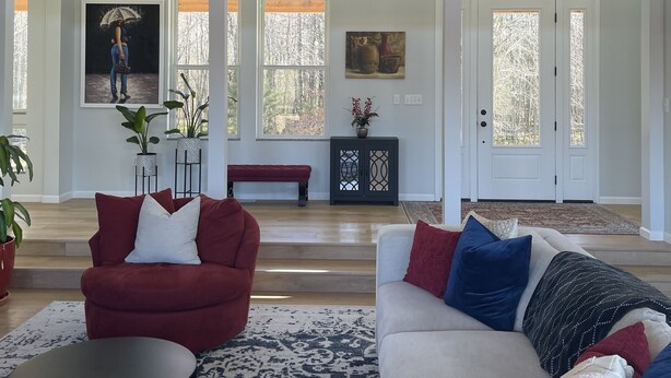 Ein geräumiges Wohnzimmer mit einem großen Kronleuchter, einem roten Sessel, einem weißen Sofa und einem schwarzen Couchtisch. In der Nähe der Fenster befinden sich Kübelpflanzen und eine Bank.