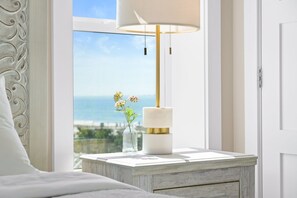 A bedroom with a bed, a nightstand with a lamp, a vase with flowers, and a view of the sea. - Pinkies Up: Stunning MB Home- 4BR, Beachfront, Pet Friendly, Amazing Views! (Mexico Beach)