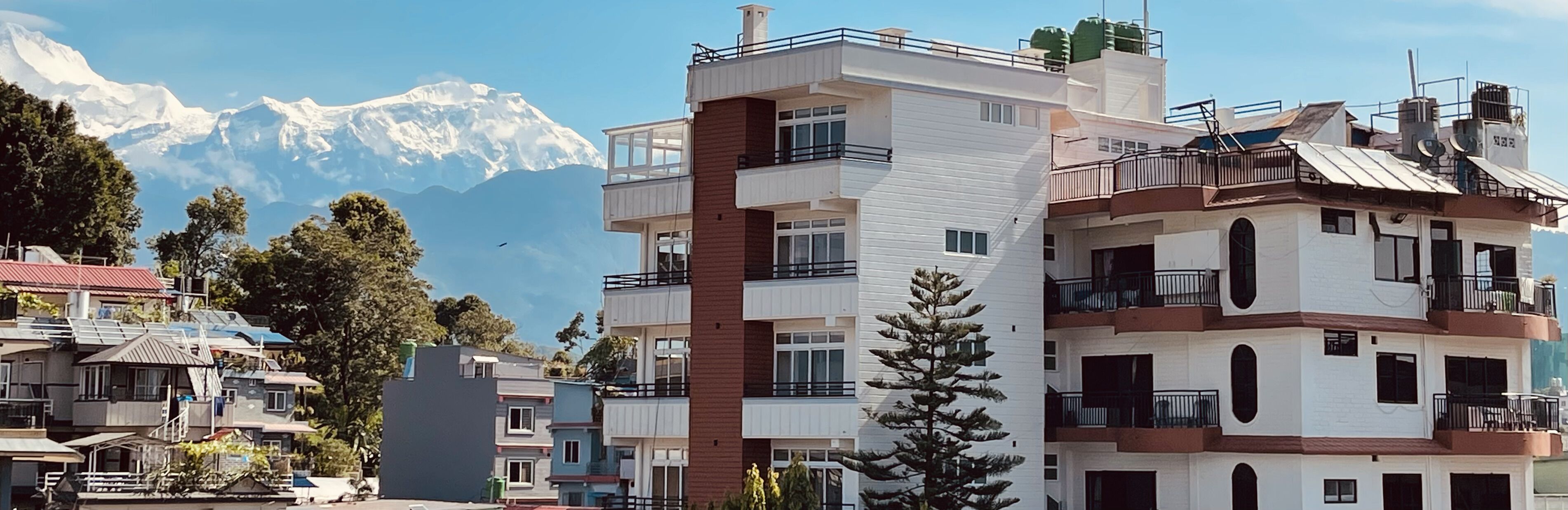 A modern multi-story building with balconies, located in a mountainous area with snow-capped peaks in the background.