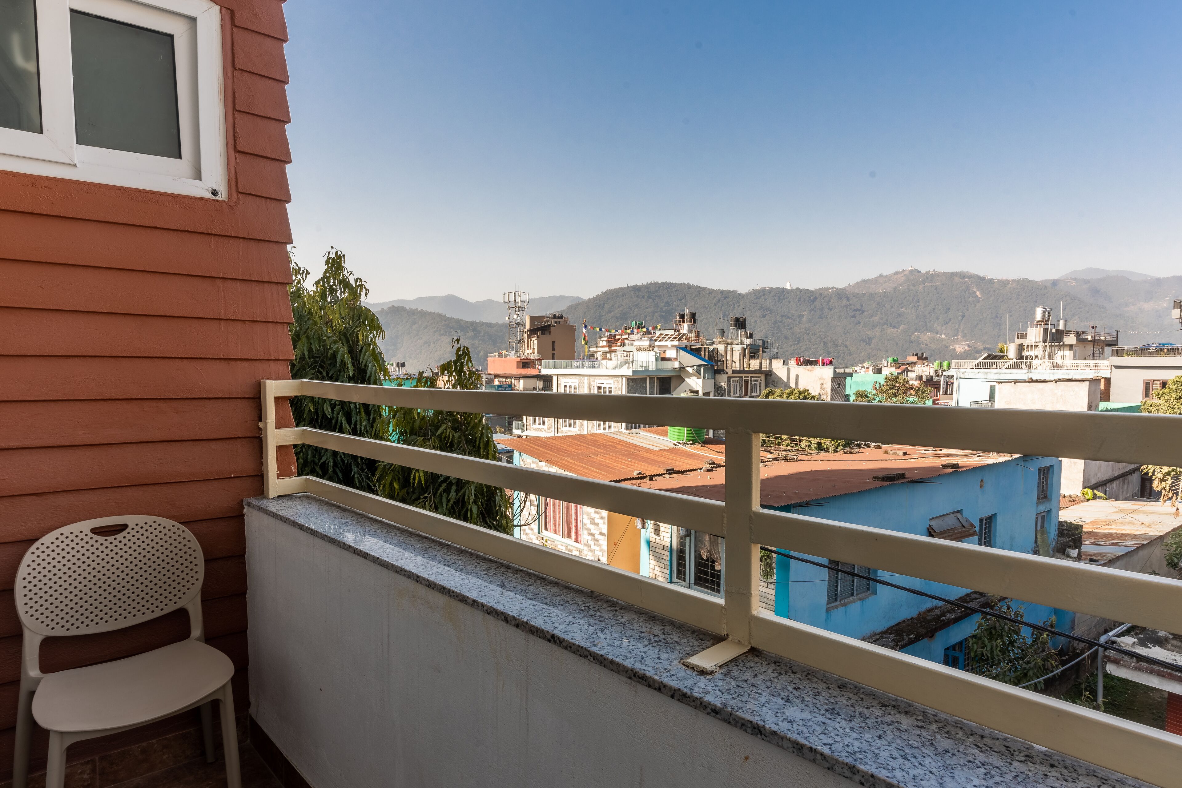 Un balcon avec une chaise blanche, d’où l’on peut admirer un paysage urbain avec des bâtiments et des montagnes en arrière-plan.