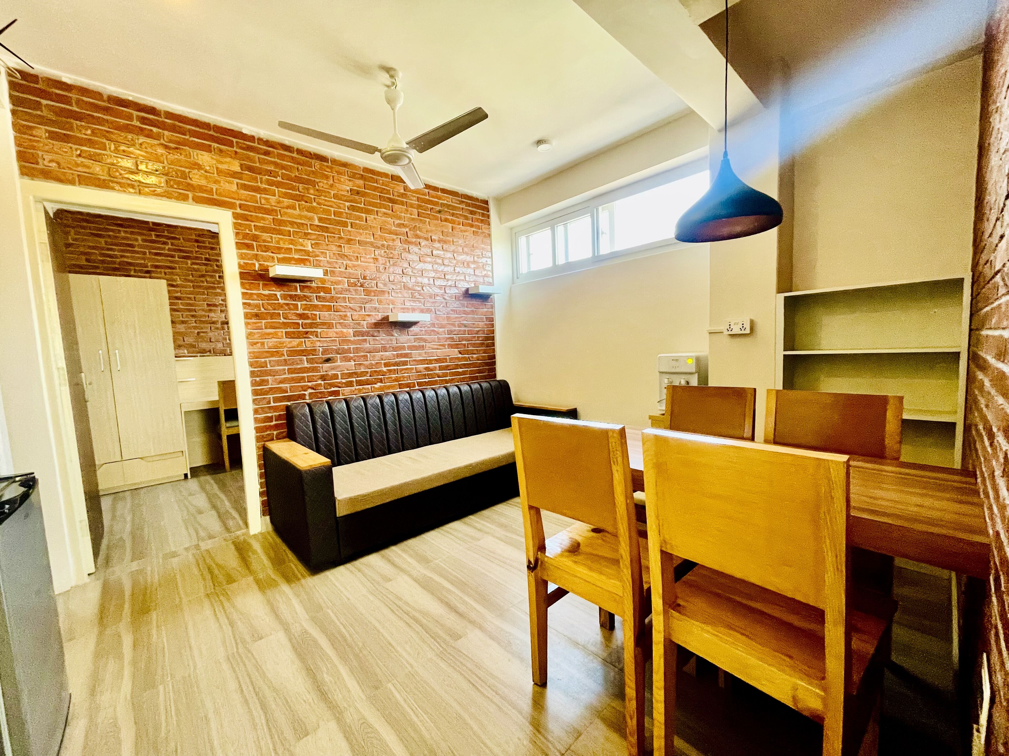 A modern living room with a brick wall, a ceiling fan, a wooden bench, and a dining area with wooden chairs and a table.
