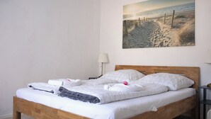 Una cama bien hecha con cabecera de madera, un cuadro colgado en la pared que muestra una escena de playa y una mesita de noche con una lámpara.