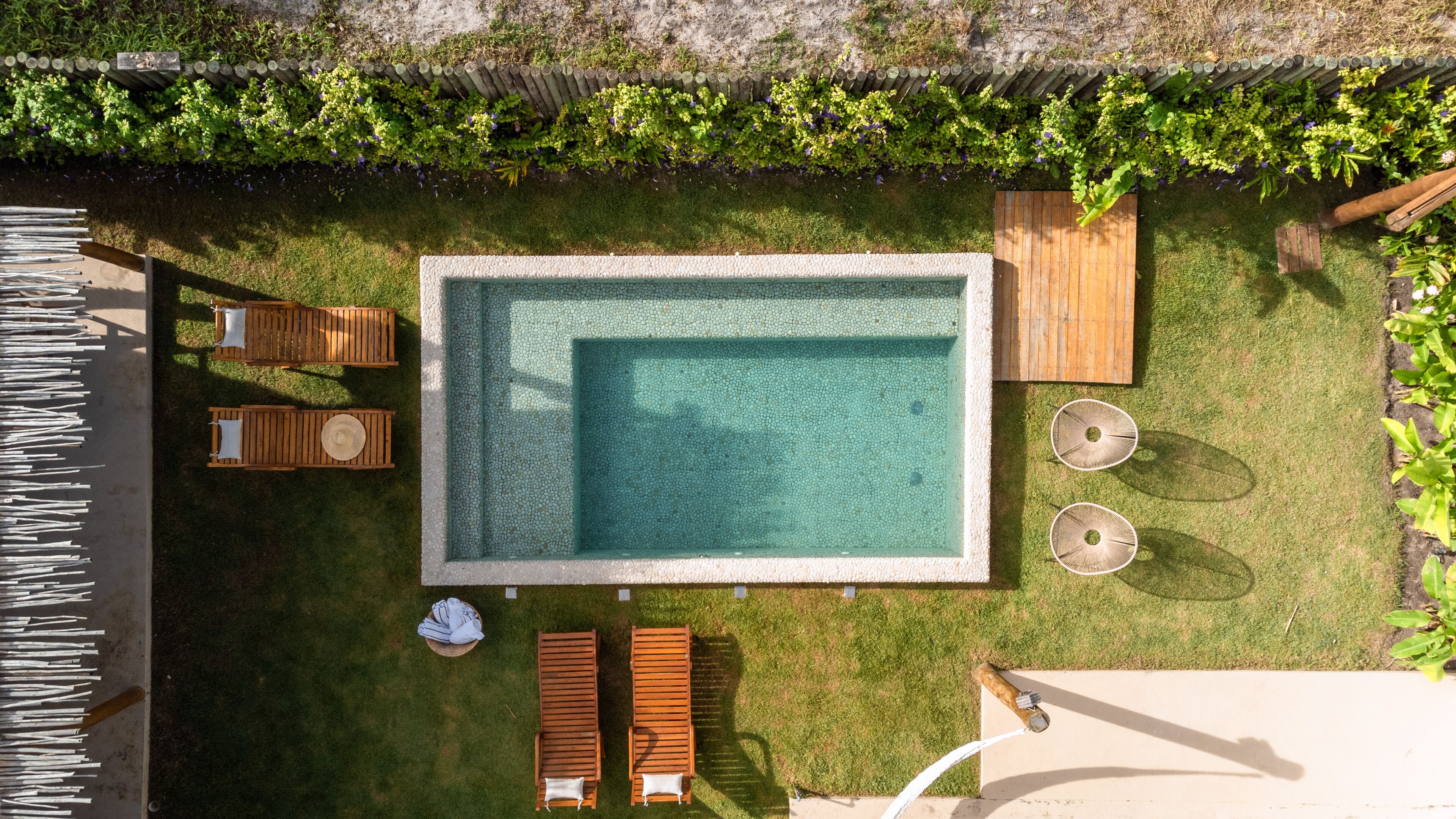 A rectangular swimming pool with a tiled deck, surrounded by a wooden deck and a grassy area. There are two wooden lounge chairs and a small table with a white cloth.