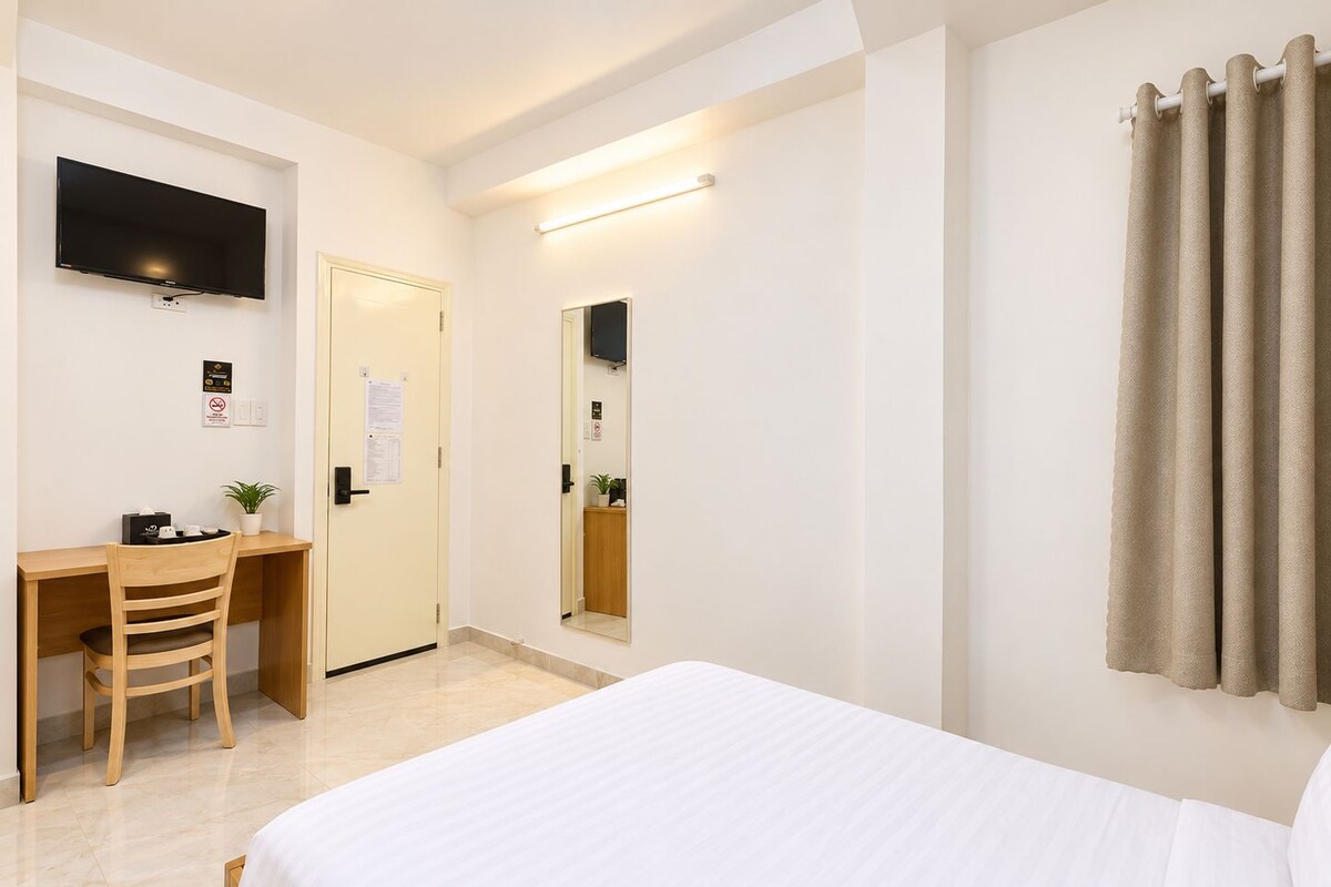 A hotel room with a bed, a desk with a chair, a flat-screen TV mounted on the wall, a mirror, and a window with curtains.