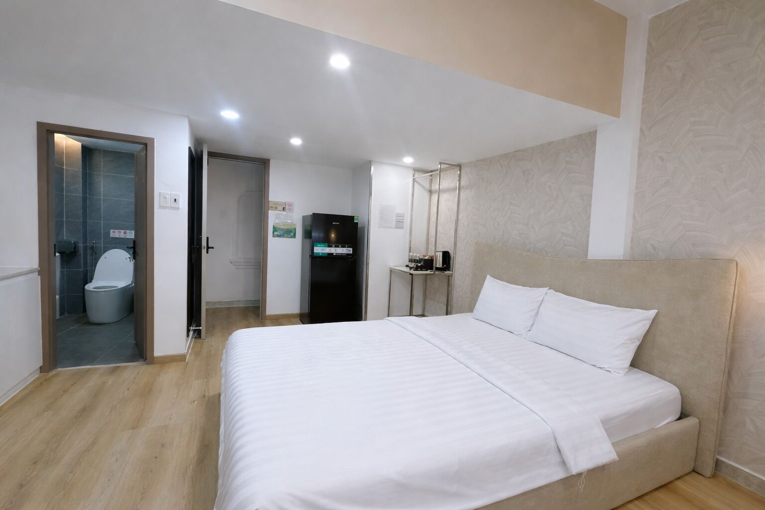 A hotel room with a bed, a bathroom with a toilet and sink, a refrigerator, and a small table with a mirror.