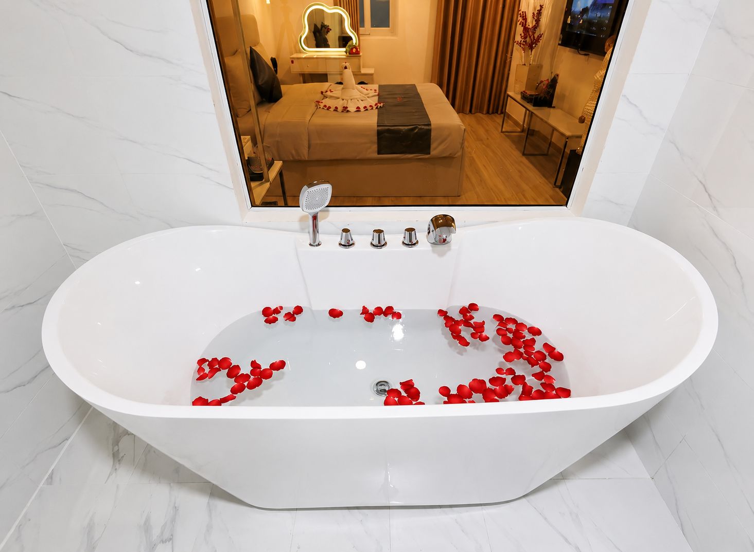 Un baño moderno con una bañera independiente llena de pétalos de rosa, un tocador con un espejo, y una cama visible en el reflejo.