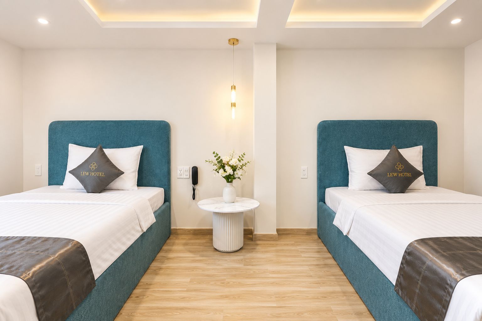 A hotel room with two beds, each with a teal headboard, white bedding, and a pillow with the hotel's logo. There's a small round table with a vase of flowers and a phone on the wall.