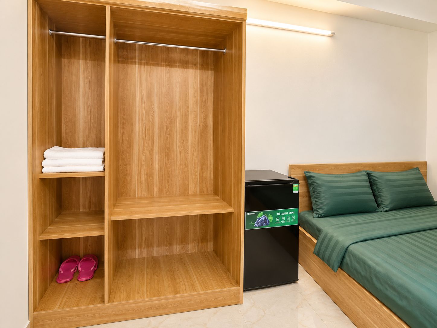 A hotel room with a wooden wardrobe, a bed with green bedding, a small black refrigerator, and pink slippers.