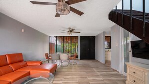 Amplia sala de estar con un sofá naranja moderno, una mesa de centro de vidrio y una mesa de comedor con sillas. Hay dos ventiladores de techo, una escalera y un televisor montado en la pared.