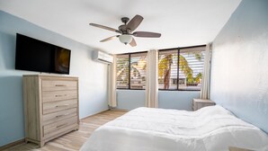 Un dormitorio con cama, ventilador de techo, televisor, cómoda y ventana con persianas.