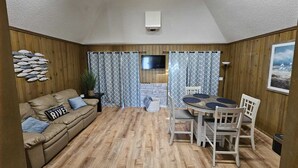 Une pièce chaleureuse avec un plancher en bois, un canapé agrémenté d’un coussin orné du motif « River », une table à manger avec des chaises et un téléviseur fixé au mur.