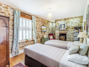 Ein Schlafzimmer mit Steinwand, einem Bett mit gestreifter Bettwäsche, einem Holzschrank, einem Fenster mit floralen Vorhängen und einer Pendelleuchte.