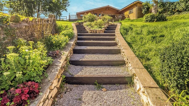 Eine Holztreppe führt zu einem Gebäude mit einer Steinverkleidung, umgeben von üppiger Vegetation und blauem Himmel.