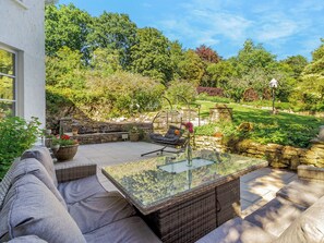 Eine Terrasse mit Glastisch und Korbmöbeln, umgeben von einer Steinmauer und üppiger Bepflanzung.
