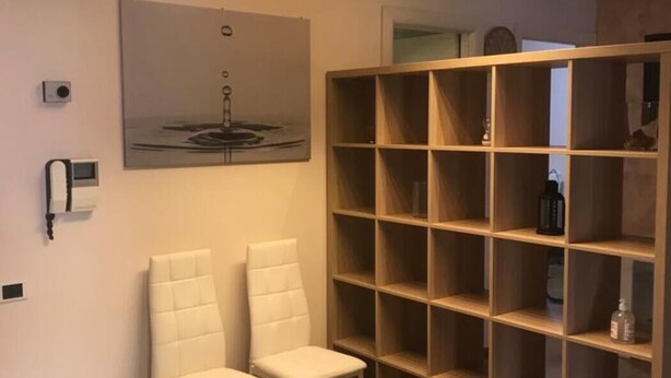 Huoneessa on kaksi valkoista tuolia, suuri puinen hyllykkö, kattovalaisin ja seinälle ripustettu taideteos.